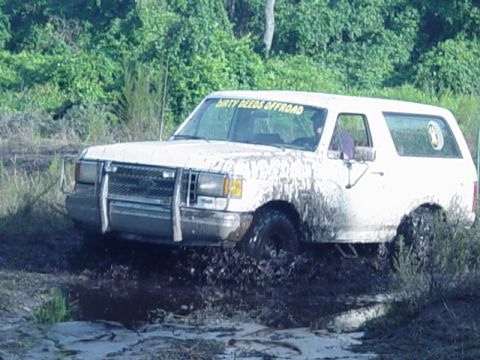 1989 Ford Bronco - White Ford Bronco stuck in mud, with 'DIRTY DEEDS OFFROAD' sticker on windshield.