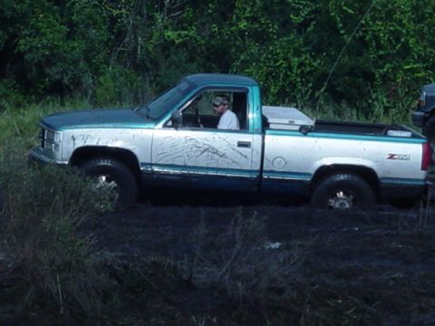 1989 Ford Bronco - A two-tone pickup truck is stuck in dark, muddy terrain with trees in the background.