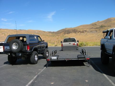 1985 Ford Bronco - A lifted black 1985 Ford Bronco is parked in a lot with a trailer and other vehicles under a clear blue sky.