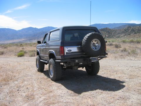 1985 Ford Bronco - Rear view of a lifted 1985 Ford Bronco with a spare tire on the back, parked in a dry, hilly landscape.