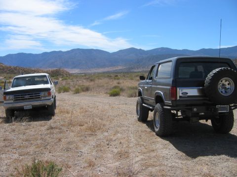 1985 Ford Bronco - Two SUVs are parked in a desert landscape under a bright blue sky with mountains in the distance.