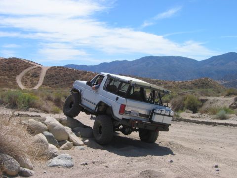 1985 Ford Bronco - Modified 1987 Ford Bronco navigates a rocky obstacle on an off-road trail under a blue sky.