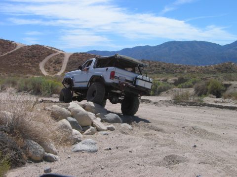 1985 Ford Bronco - This modified SUV is shown tackling a rocky obstacle on a desert trail under a partly cloudy sky.
