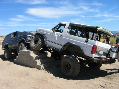 1985 Ford Bronco - Two modified Ford Broncos tackle an obstacle course on a sandy terrain under a blue sky.