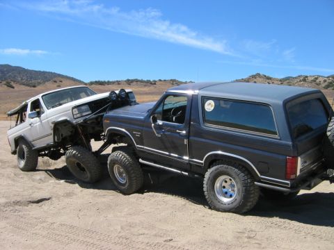 1985 Ford Bronco - Two heavily modified SUVs are seen off-roading on a sunny day with hills in the background.