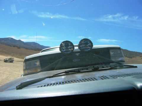 1985 Ford Bronco - View from inside a vehicle shows a 1985 Ford Bronco with auxiliary lights, parked in a dry, open landscape.