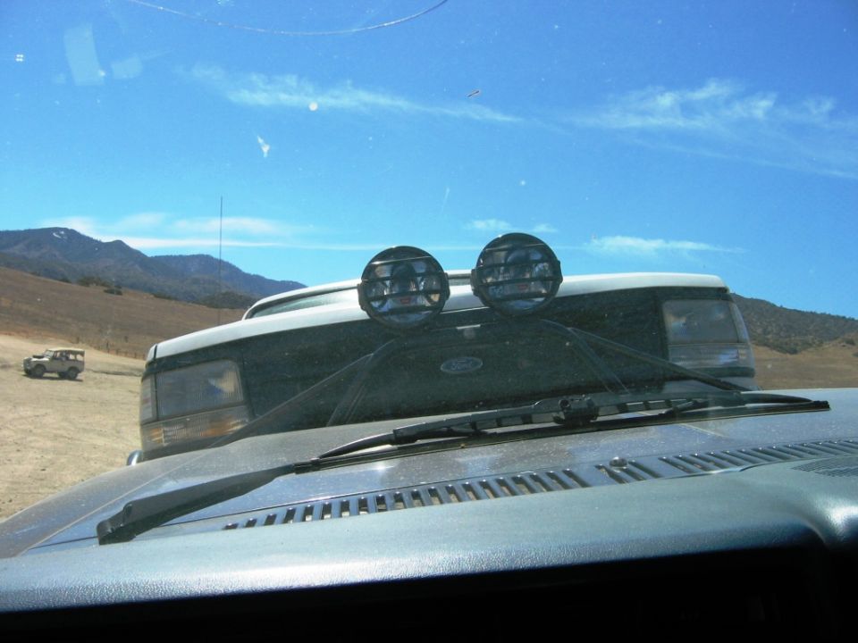 Windshield view over dashboard showing hood with Ford badge and two round off-road lights, dry hills beyond