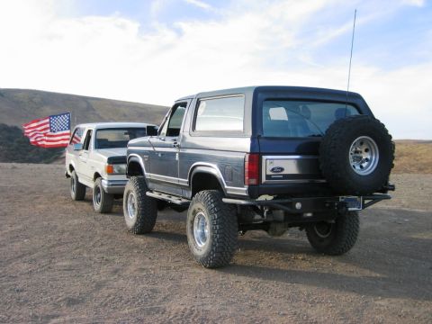 1985 Ford Bronco - Two Ford Broncos are parked on a dirt road under a cloudy sky, with an American flag visible.