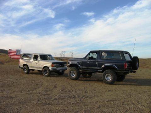 1985 Ford Bronco - Two Ford Broncos are parked on a dirt lot under a cloudy sky, with power lines in the background.