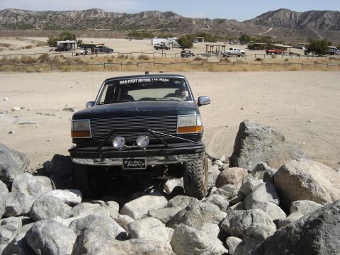 1987 Ford F-250 - A dark-colored Ford Bronco is shown driving over rocks in a desert-like environment with mountains in the background.