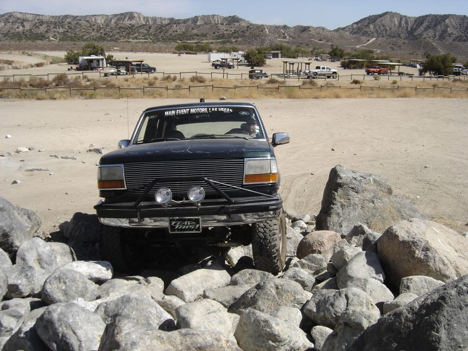 Ford Bronco navigating rocky terrain. Has 'Main Event Motors Las Vegas' on windshield banner and 'Team Trust' sticker on bumper