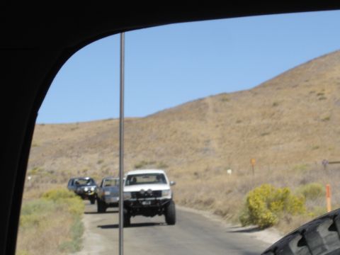 1987 Ford F-250 - A convoy of trucks, including a modified white Ford, travels on a dusty road through a dry, hilly landscape.