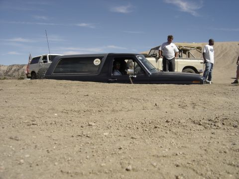 1987 Ford F-250 - A dark SUV is partially buried in sand with a white pickup truck visible in the background.