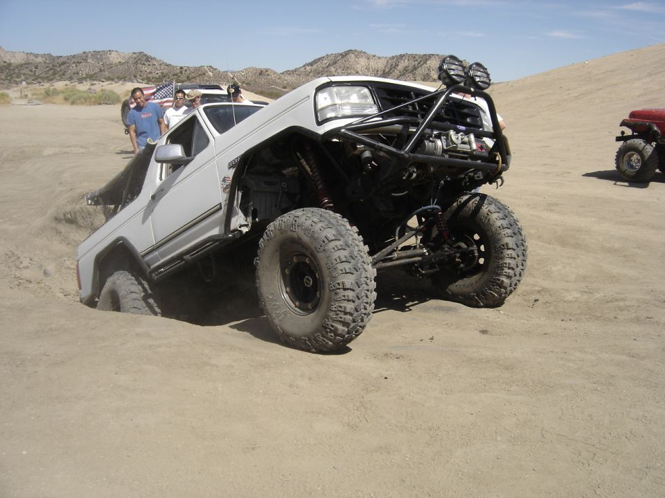 White off-road truck with custom roll cage stuck in sand, large tires visible