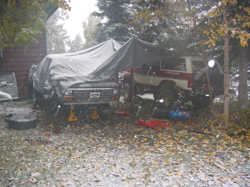 1980 Ford Bronco front end under tarp, another Bronco visible behind it, snow falling