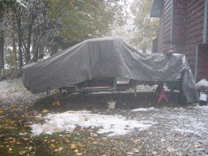 Tarp-covered truck body on jack stands in a snowy yard during falling snow