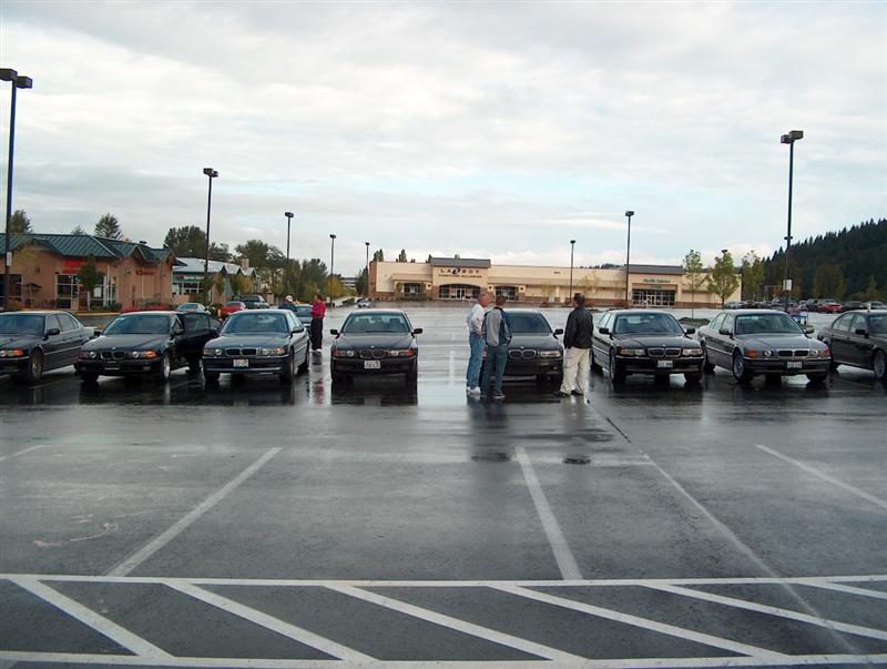 Row of dark BMW sedans parked in a wet lot with people gathered near a retail building