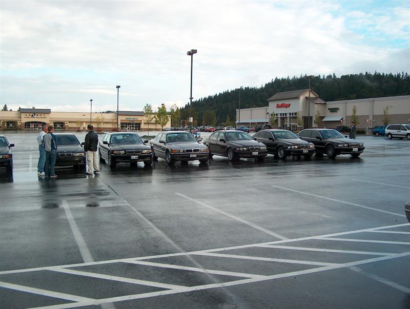 Six dark BMW sedans parked in a row at a wet shopping-center parking lot during a car meet