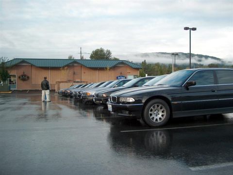 1999 BMW 7 Series - A line of black BMW 7 Series sedans are parked in a lot on a cloudy, wet day, with one car prominently in the foreground.