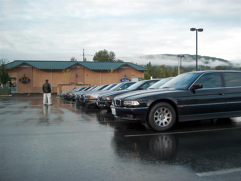 Black BMW sedan front three-quarter with a row of dark BMWs in a wet parking lot