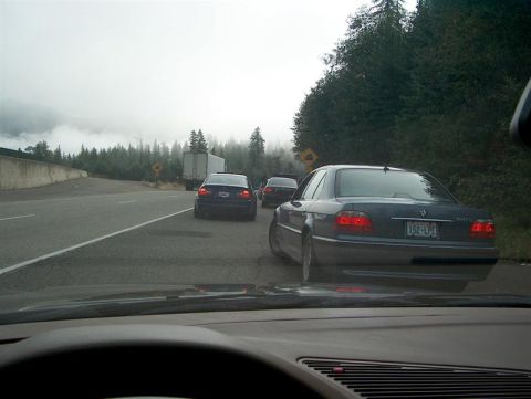 1999 BMW 7 Series - A convoy of BMW sedans travels down a misty highway, with a truck visible in the distance.