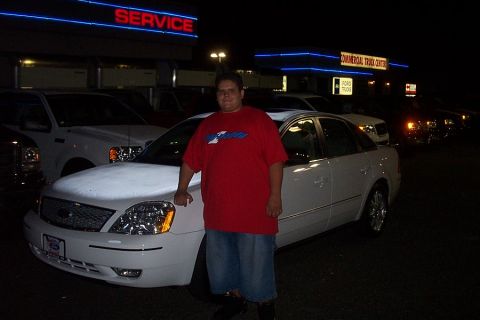 1993 Ford Taurus - Man poses with a white sedan in front of a commercial truck center at night, with other vehicles in the background.