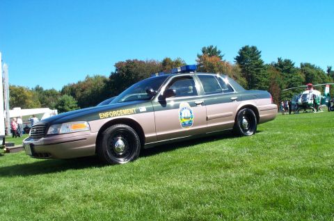 1991 Ford LTD Crown Victoria - A two-tone police car with "ENFORCEMENT" text and a light bar is parked on a grassy field with a helicopter in the background.