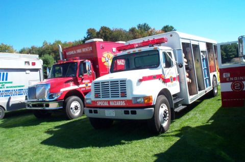 1991 Ford LTD Crown Victoria - Two fire rescue trucks, one red and one white, are parked side-by-side on a grassy field under a clear blue sky.