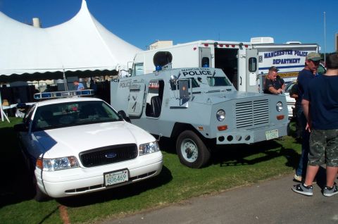 1991 Ford LTD Crown Victoria - A white police car and a gray armored police vehicle are displayed outdoors under a tent and clear blue sky.