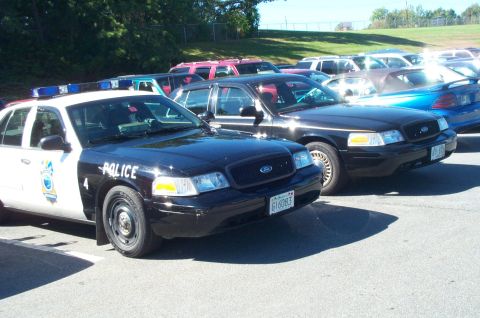 1991 Ford LTD Crown Victoria - Several police vehicles are parked in a lot on a sunny day, with trees and a fence in the background.