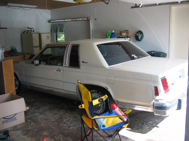 Cream two-door Ford LTD in a garage on jack stands with wheel off, rear 3/4 view