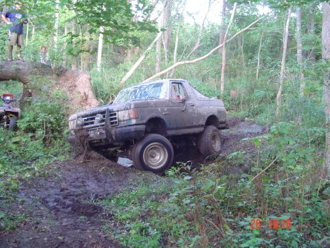 1989 Ford Bronco - A muddy Ford Bronco navigates a challenging off-road trail with dense woods on all sides.