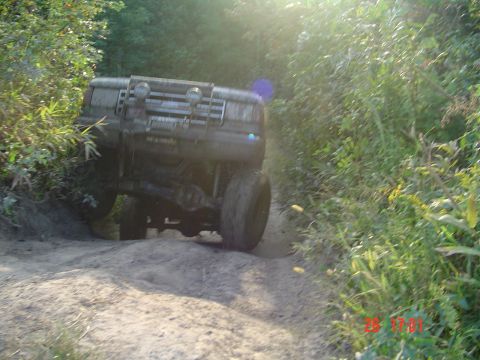 1989 Ford Bronco - A muddy 1987 Ford Bronco navigates a challenging off-road trail with dense foliage on either side.