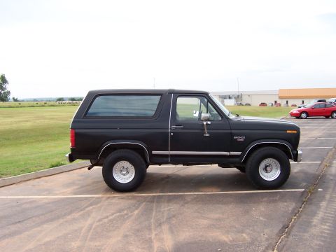 1986 Ford Bronco - This black Ford Bronco is parked in a lot with a red car visible in the background.