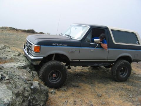 1989 Ford Bronco - This lifted Ford Bronco navigates a rocky incline on a dry, dusty landscape under an overcast sky.