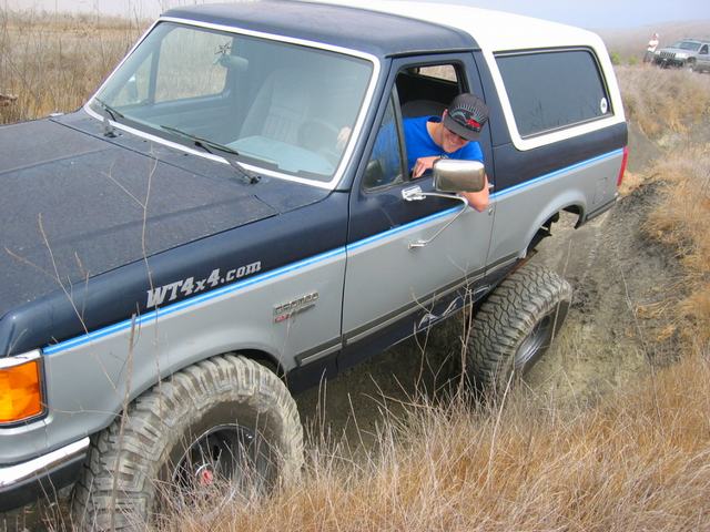 Blue and silver Ford Bronco with white top on a rutted dirt trail, driver leaning out the window
