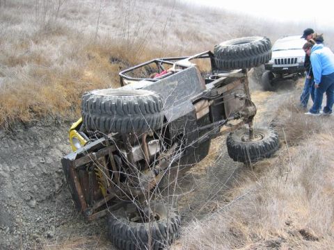 1989 Ford Bronco - An off-road vehicle is overturned on a dirt track, with a white Jeep and onlookers nearby.