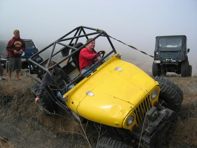 Yellow caged Jeep buggy tipped in a dirt rut on a foggy hill with a recovery strap