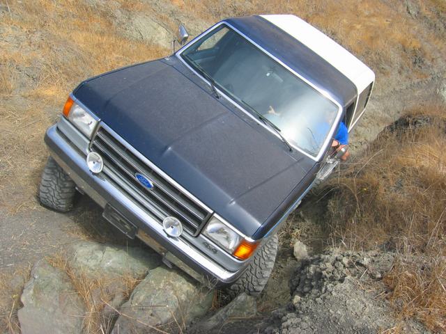 Overhead front view of a dark blue Ford Bronco with white top tipped in a dirt rut off-road