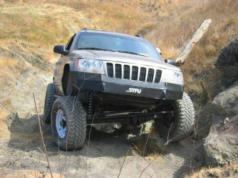 1989 Ford Bronco - This modified Jeep Grand Cherokee is shown off-roading on a dirt path with large rocks and dry vegetation.