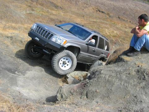 1989 Ford Bronco - Jeep Grand Cherokee tackles a challenging off-road trail with a spectator watching from the side.