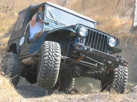 1989 Ford Bronco - A dark green Jeep CJ-5 navigates a rocky off-road path, kicking up dust.