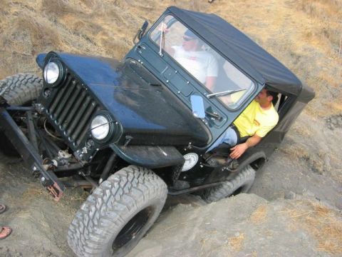 1989 Ford Bronco - A dark green Jeep CJ-7 is shown off-roading on a steep dirt hill, with two men inside the vehicle.