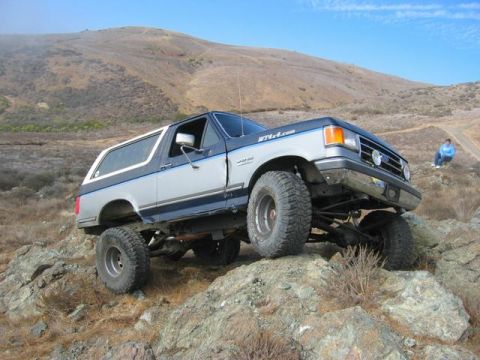 1989 Ford Bronco - A lifted Ford Bronco navigates a rocky, off-road trail under a clear blue sky.