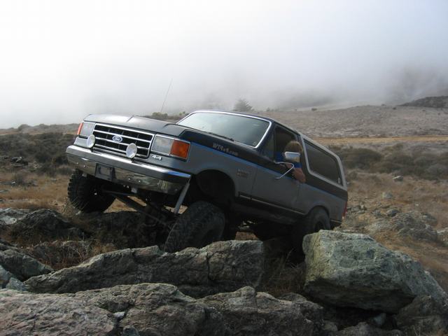 Ford Bronco climbing over large rocks with fog in the background. The vehicle has a two-tone paint scheme