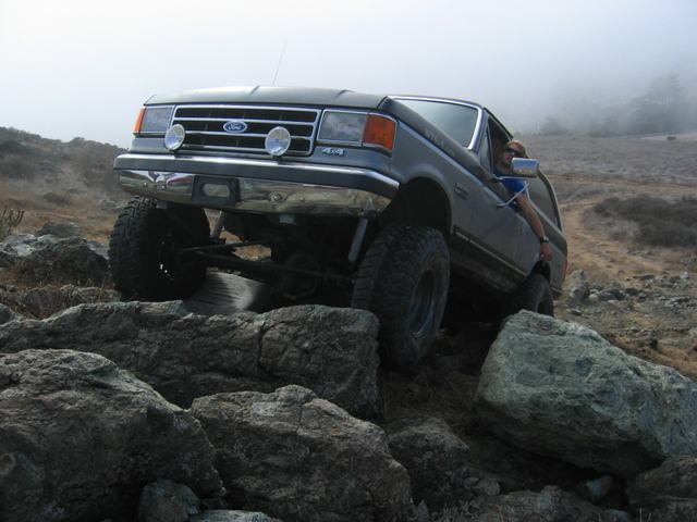 Dark gray Ford Bronco climbing over rocks, driver visible, fog in background