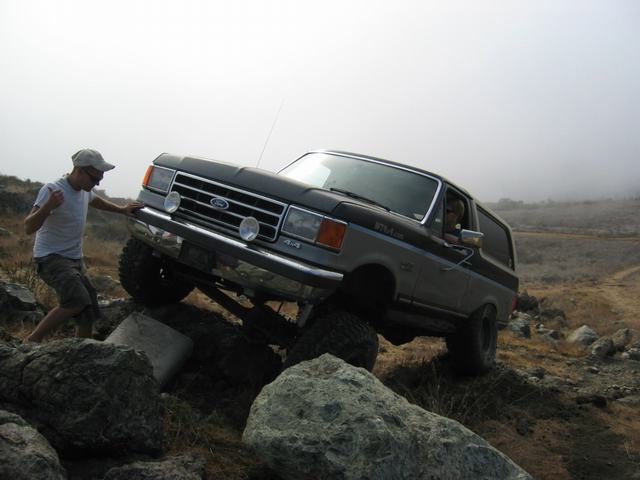 Two-tone gray Ford Bronco climbing boulders in fog, front three-quarter view, spotter alongside