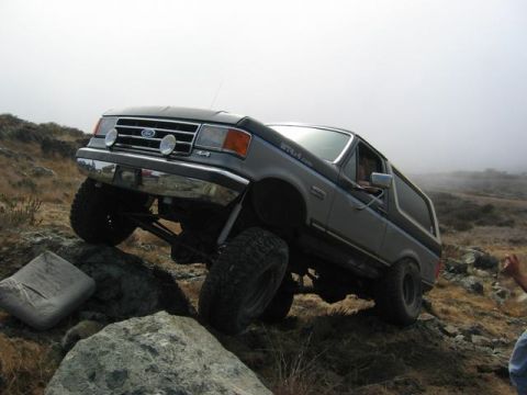 1989 Ford Bronco - A lifted Ford Bronco navigates a rocky, off-road trail under a hazy sky.
