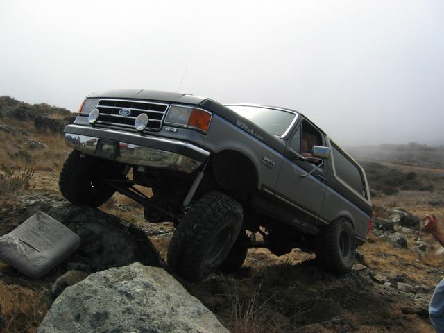 Gray Ford Bronco lifted on mud tires climbing a rock ledge in fog