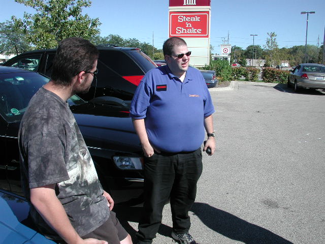 Two men stand in a parking lot near a black SUV and a blue car. A Steak 'n Shake sign is visible
